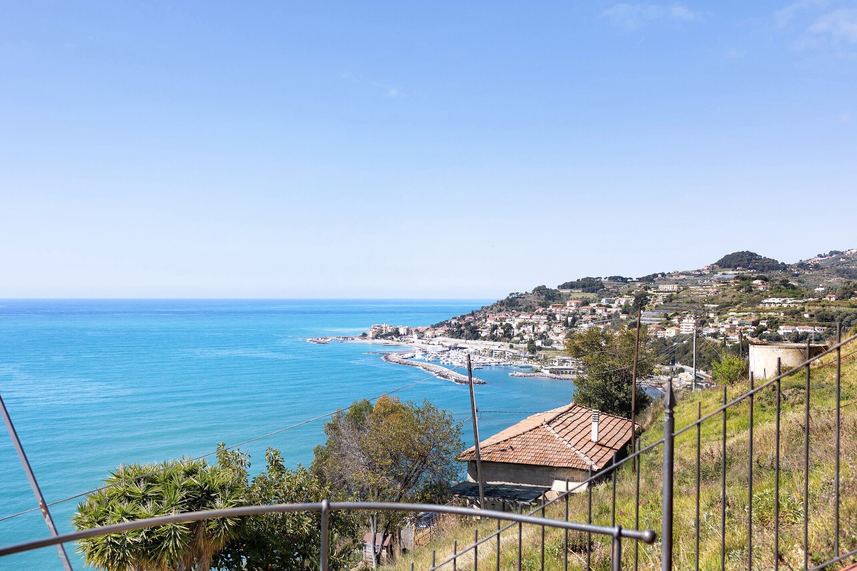 A coastal town with a clear view of the sea, a small building with a tiled roof, and a hilly landscape in the background.