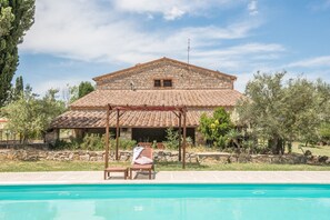 A stone building with a tiled roof, a pool, and lounge chairs.