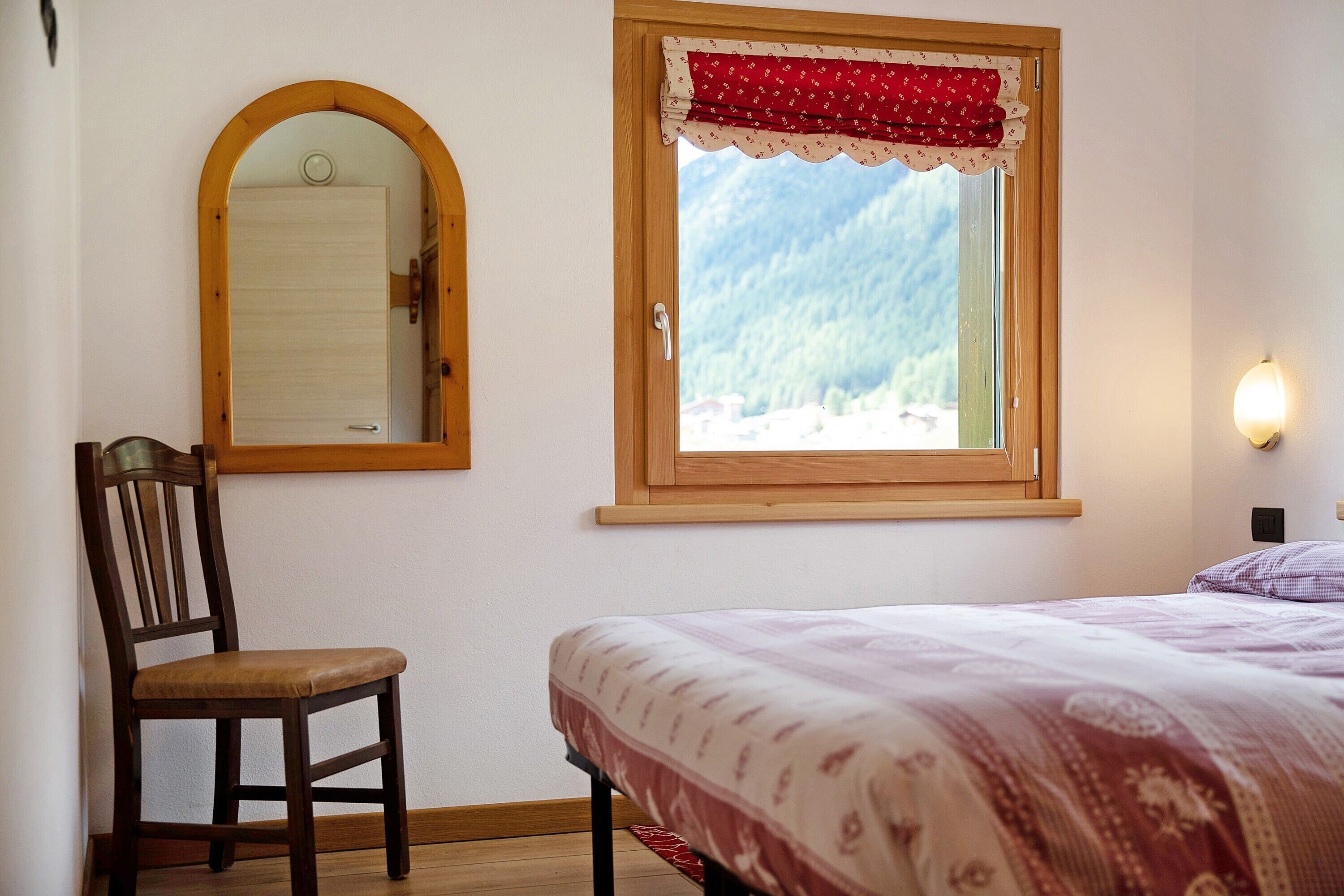 A bedroom with a bed, a chair, a window with a view of mountains, and a mirror on the wall.