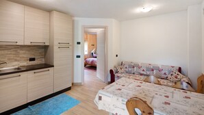 A compact kitchen with modern cabinetry, a sink, and a blue rug. A bed with patterned bedding is visible in the adjacent room.
