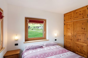 Camera da letto con finestra, armadio in legno, comodino e letto con biancheria a motivi decorativi.