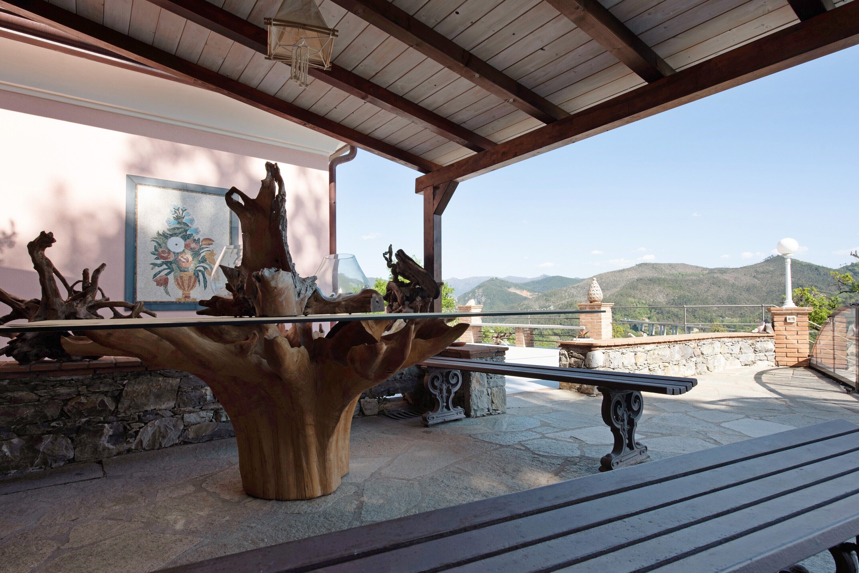 A wooden sculpture centerpiece, stone benches, and a stone wall with a painting in a covered outdoor area with a mountain view.