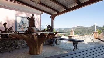 A wooden sculpture centerpiece, stone benches, and a stone wall with a painting in a covered outdoor area with a mountain view.