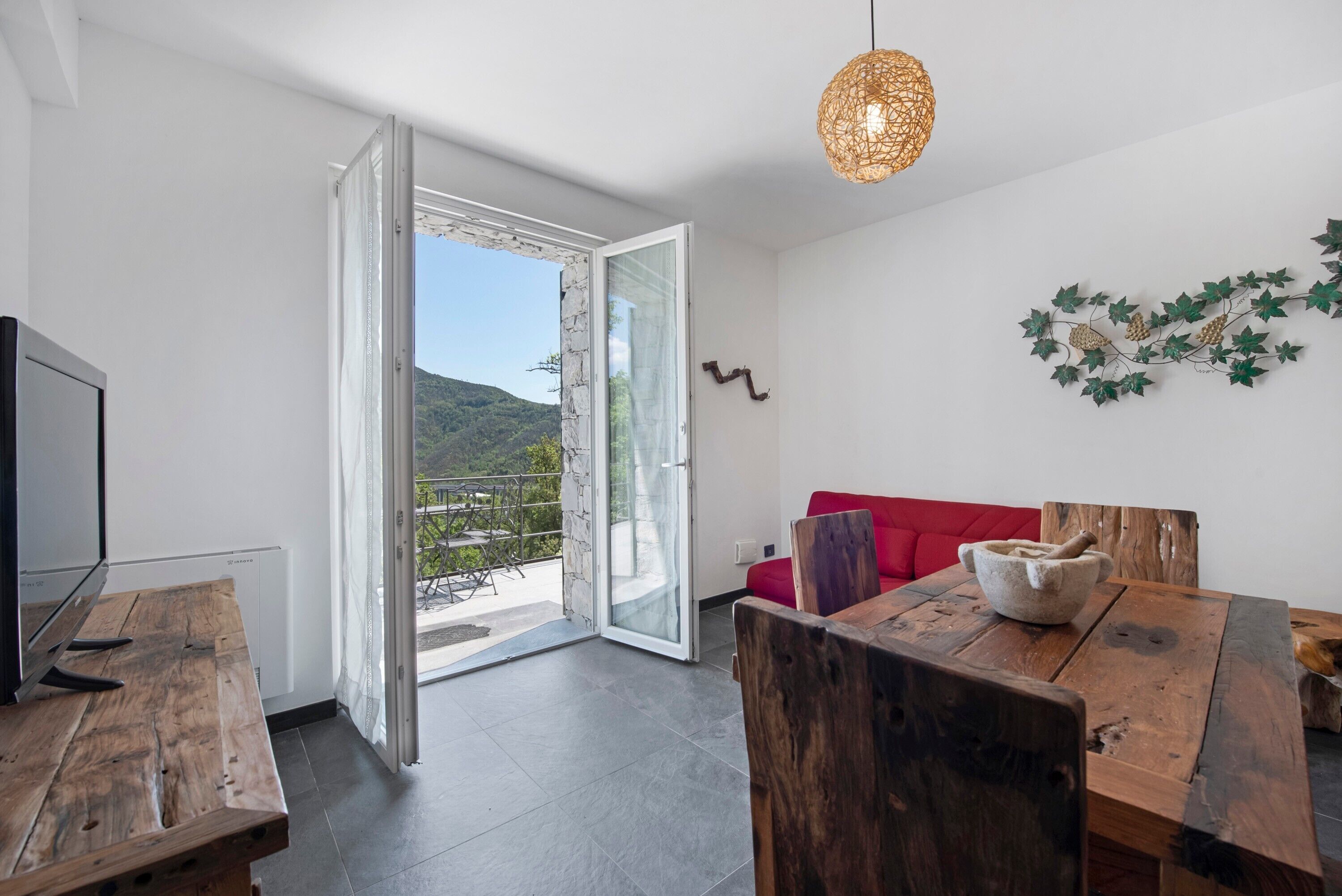 Una stanza con un tavolo da pranzo in legno, un divano rosso, una televisione e vista su un paesaggio montuoso attraverso una porta aperta.