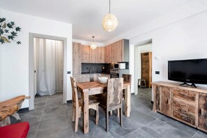 A modern kitchen with wooden cabinets, a dining table with chairs, a television on a wooden cabinet, and a hanging light fixture.