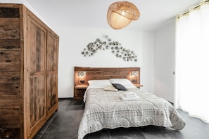 A bedroom with a wooden wardrobe, a bed with patterned bedding, a wall-mounted light fixture, and a decorative wall piece.