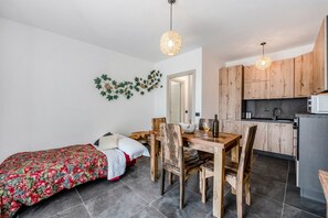 A compact room with a bed, a wooden dining table, chairs, a kitchen area with wooden cabinets, and a decorative wall piece.