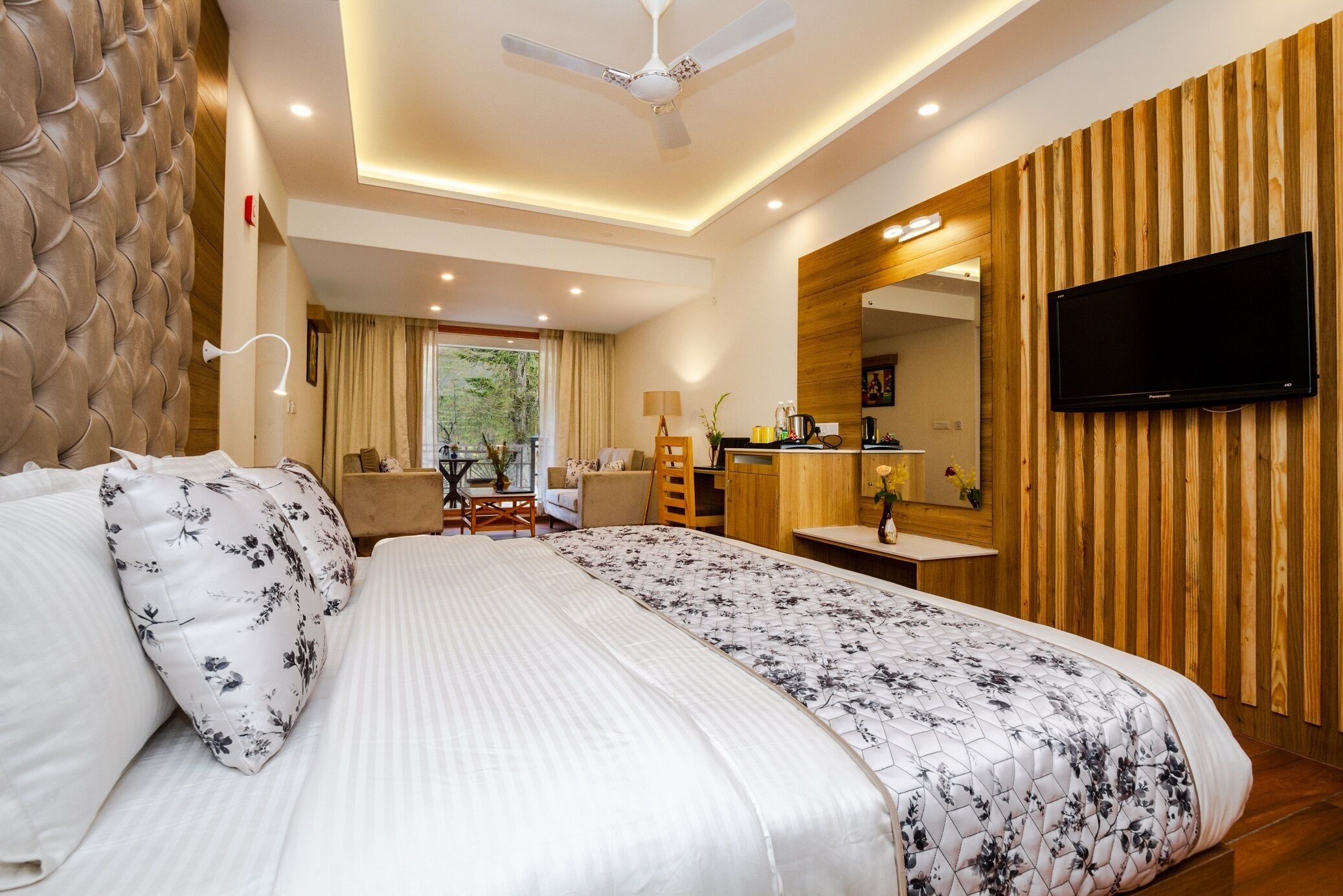 A hotel room with a large bed, a TV mounted on the wall, a wooden accent wall, and a view of the outdoors.