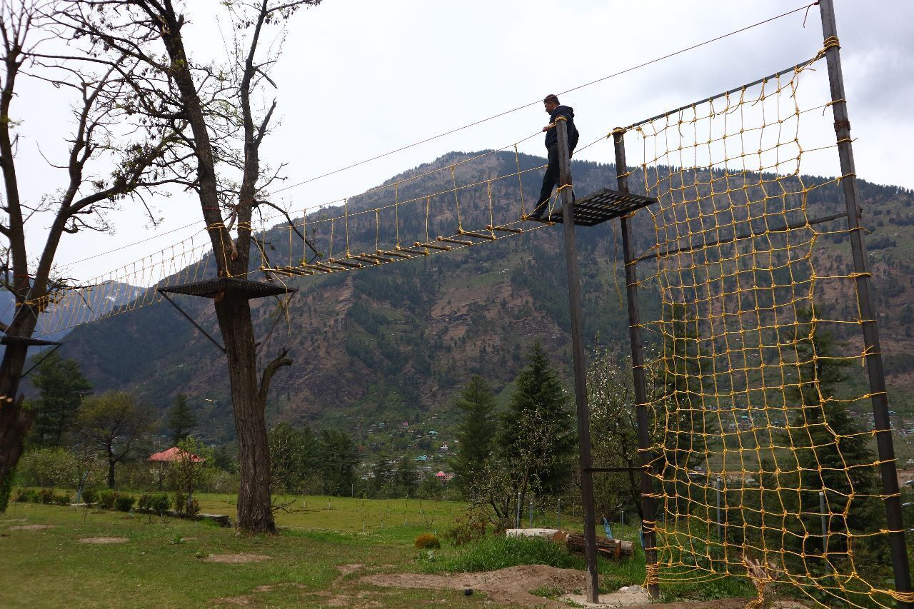 A person on a rope bridge in a mountainous area with trees and a village in the background.
