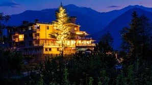 A multi-story building with balconies, a lit Christmas tree, and mountains in the background. - Anand Vardhan Resorts (Kullu)
