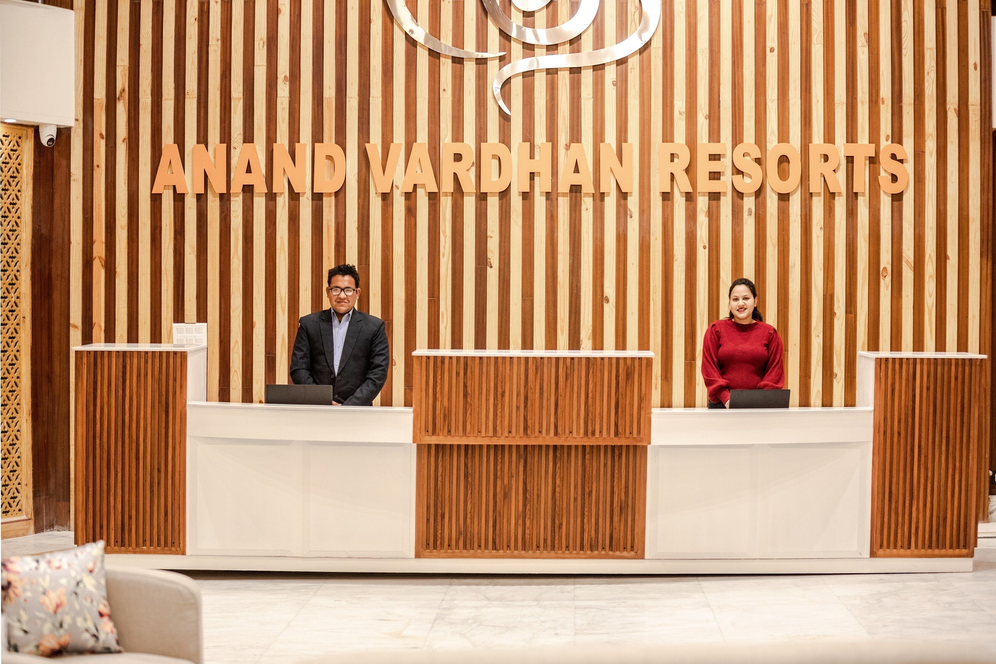 Un mostrador de recepción en Anand Vardhan Resorts, con un fondo de madera y dos empleados.