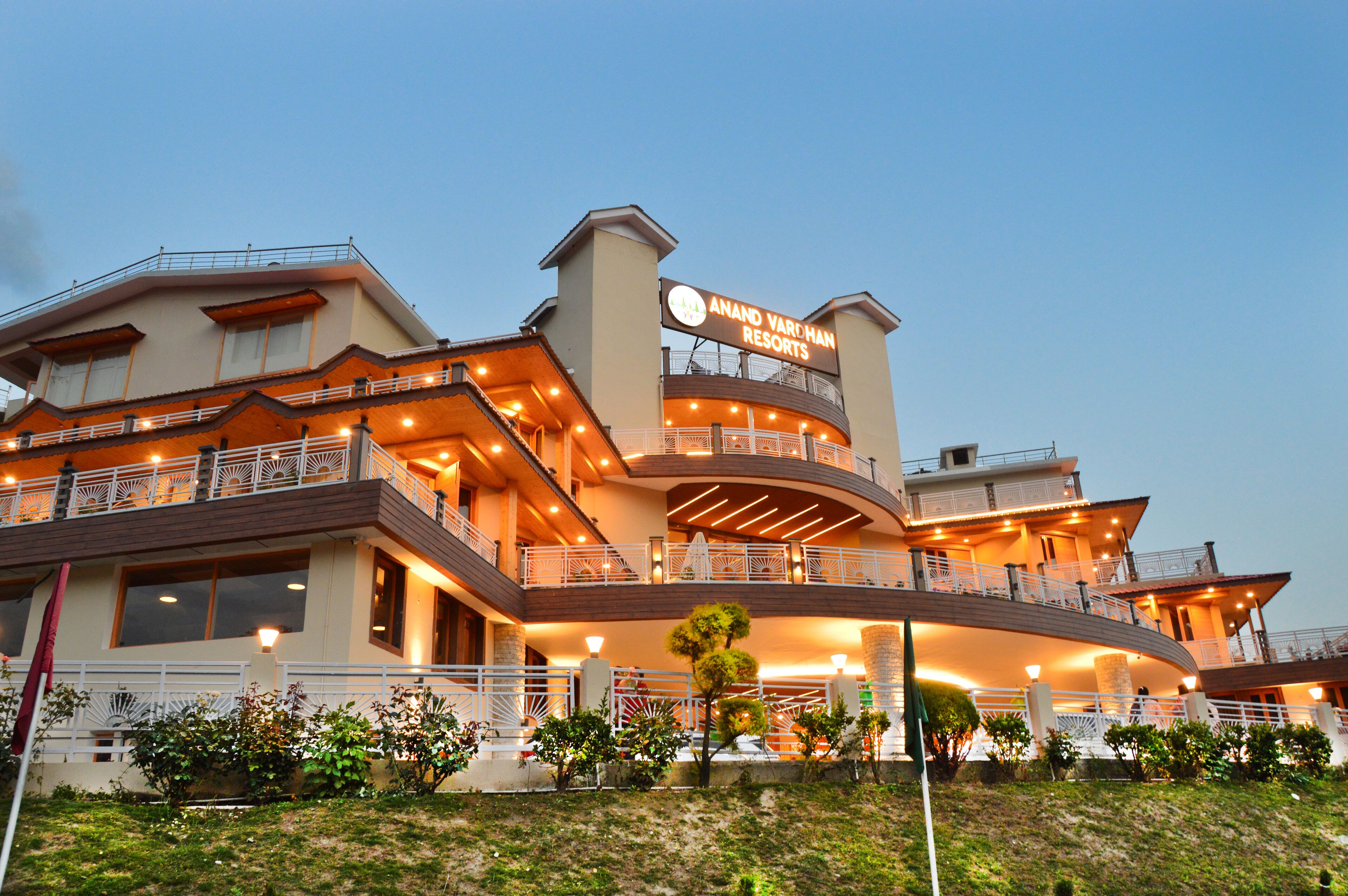 Un hotel de varios pisos con un letrero destacado que dice "Anand Varsham Resorts". El edificio cuenta con balcones, un techo curvo e iluminación cálida.
