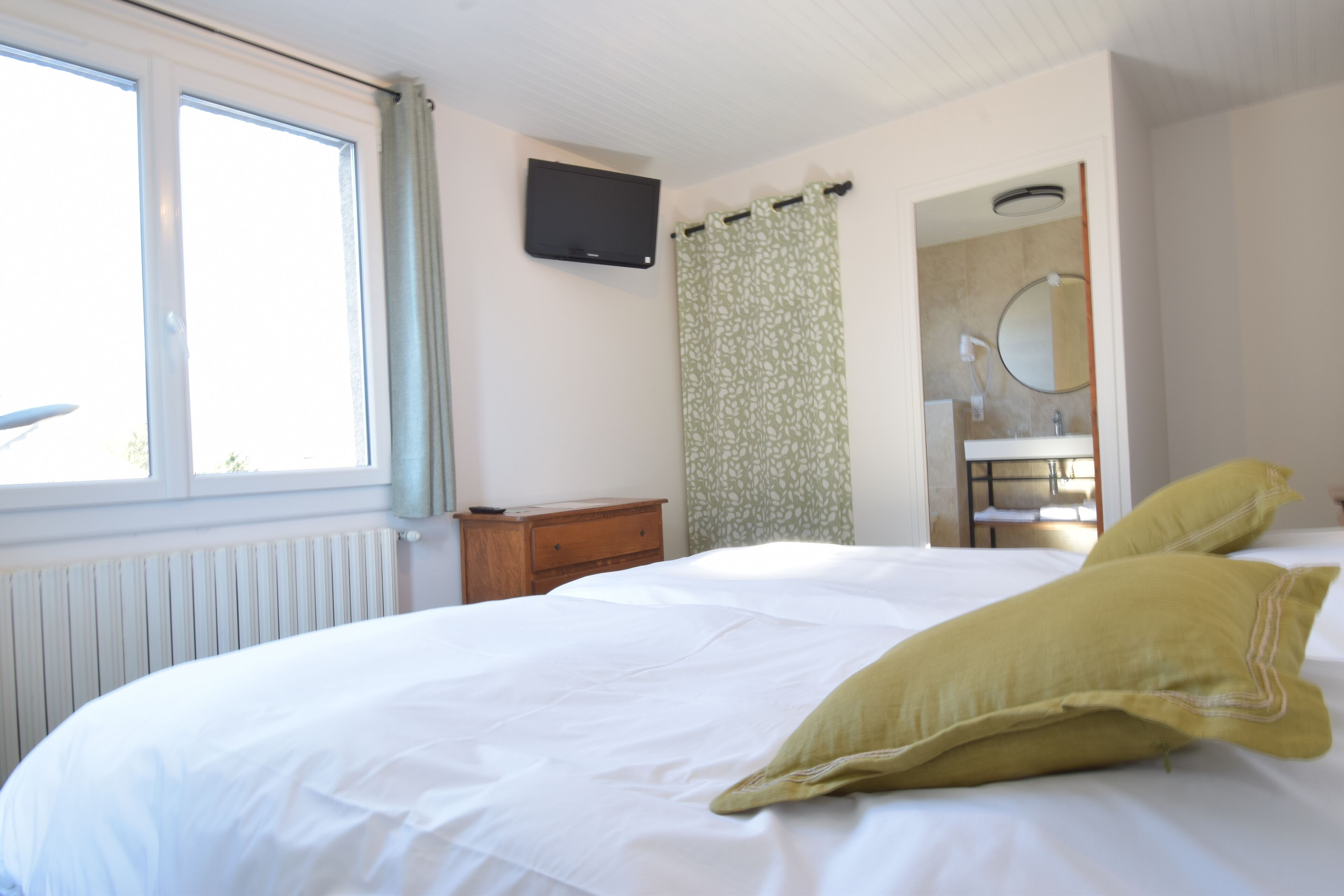 Een slaapkamer met een bed, nachtkastje, een aan de muur bevestigde televisie, een raam met gordijnen, en een badkamer met spiegel en wastafel.