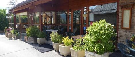 A restaurant with a covered outdoor seating area, potted plants, and a brick wall.