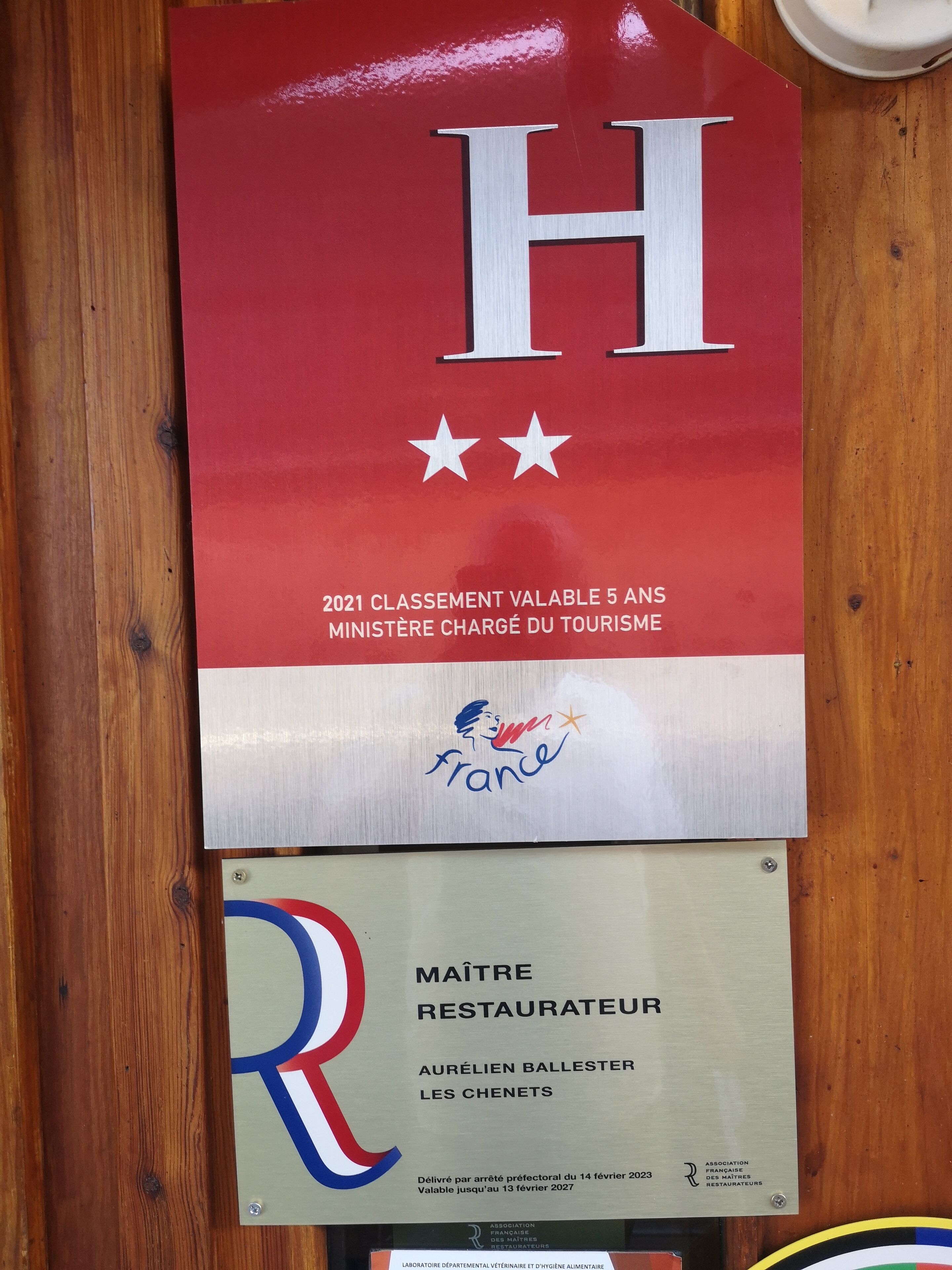 Placa com a letra 'H' e estrelas, indicando um hotel. Abaixo, placa de 'Maitre Restaurateur' Aurélien Ballester com o endereço 'Les Chenets'.