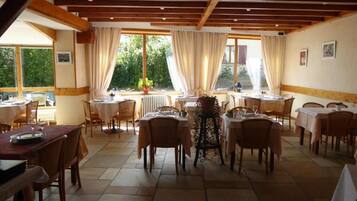 A dining area with multiple tables and chairs, large windows with curtains, and a tiled floor.