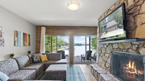 A living room with a stone fireplace, a large flat-screen TV mounted on a stone wall, a sectional sofa, and a view of a lake through large glass doors. - Carolina Sunrise by SoCharm | Amazing Sunrises | Hot tub (Denver)