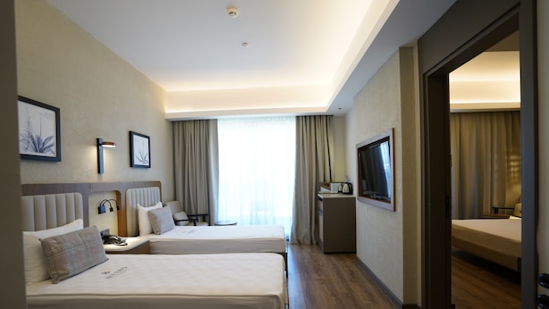 A hotel room with two beds, a desk, a TV, and a large window with curtains. - Aletris Deluxe Hotel & Spa (Manavgat)