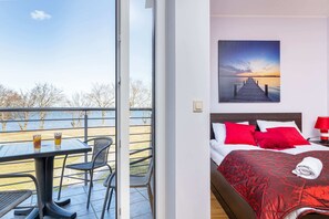 Hotellihuone, jossa on sänky, kuva laiturista ja parveke, jossa on pöytä ja tuolit.