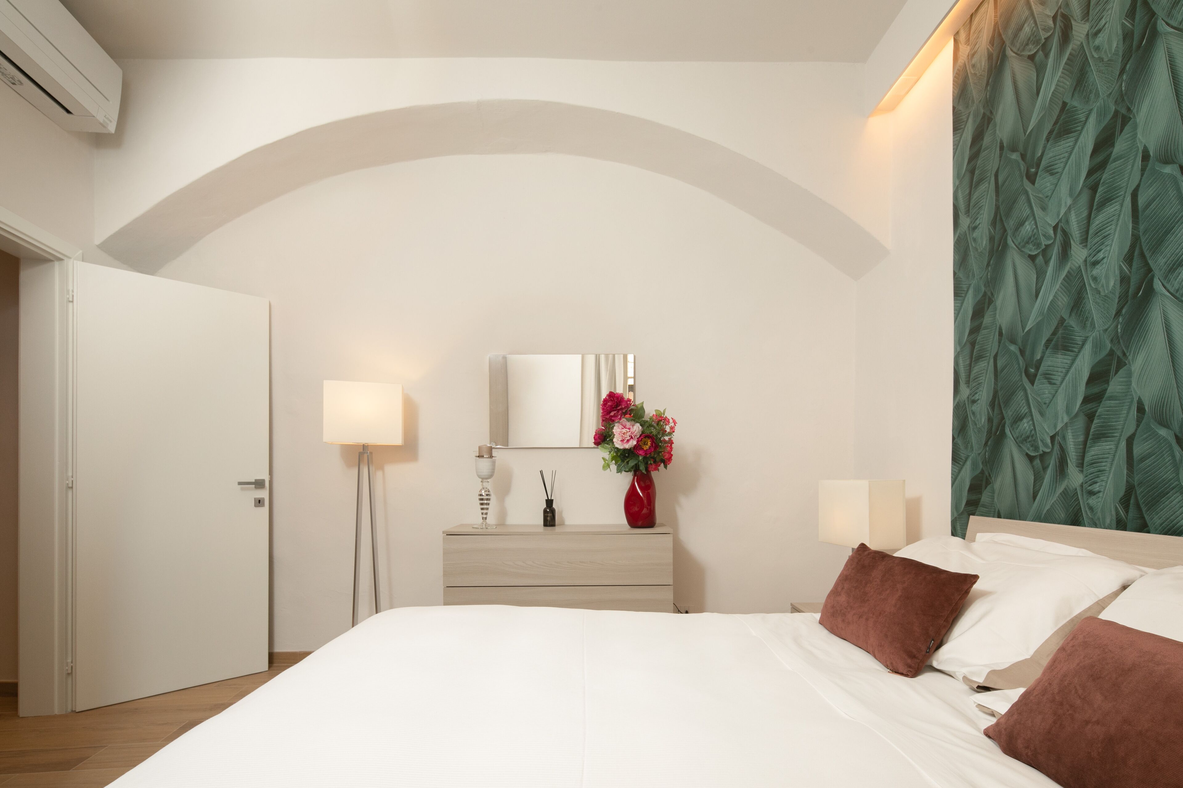 A bedroom with a bed, bedside lamps, a dresser, a mirror, and a floral arrangement.