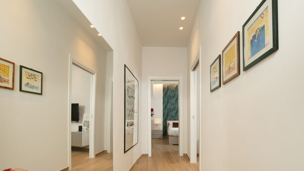 A hallway with framed artwork on the walls, a wooden floor, and a door leading to a room with a bed and a nightstand.