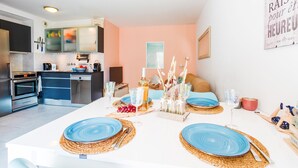 A modern kitchen with stainless steel appliances, a dining area with blue plates and a white table, and a cozy seating area with a sofa and cushions. - PALAIS LIBERTE V AP4267 By Riviera Holiday Homes (Niza)