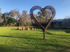 Una escultura metálica en forma de corazón en un campo cubierto de césped, con mesas de picnic y árboles al fondo.