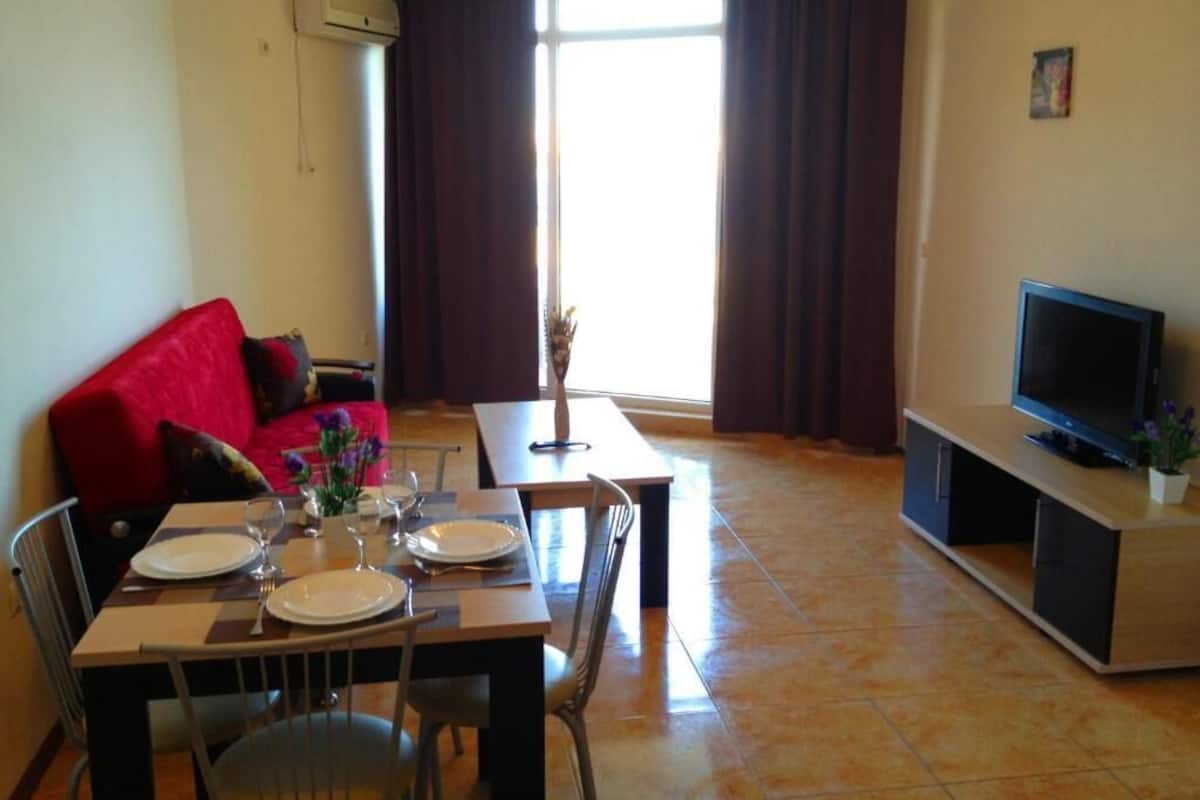 Una sala de estar con una mesa de comedor lista para dos personas, un sofá rojo, un televisor sobre un mueble bajo y un ventanal con cortinas.