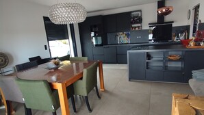 A modern kitchen with dark cabinetry, a central island, and a dining area with a wooden table and green chairs. - Villa Moderne 5 Min Mer - Piscine Idéale Familles, Proche Station de Ski 🏖️❄️ (Ventiseri)