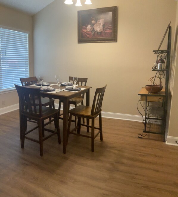 Uma sala de jantar com uma mesa de madeira posta para quatro pessoas, cercada por cadeiras. Um quadro está pendurado na parede acima da mesa.