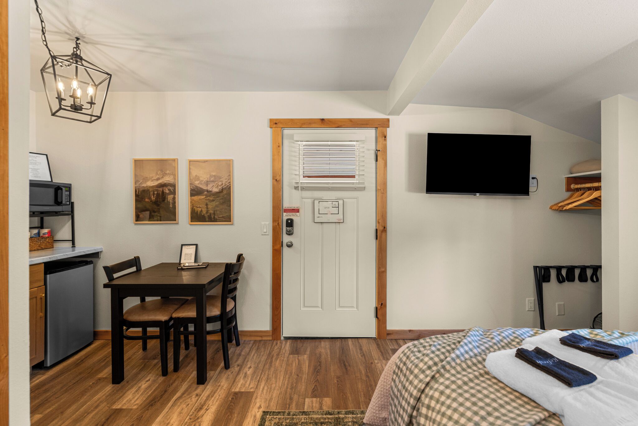 Una camera compatta con un letto, una scrivania con una sedia, un angolo cottura, una porta con finestra e una televisione a muro.