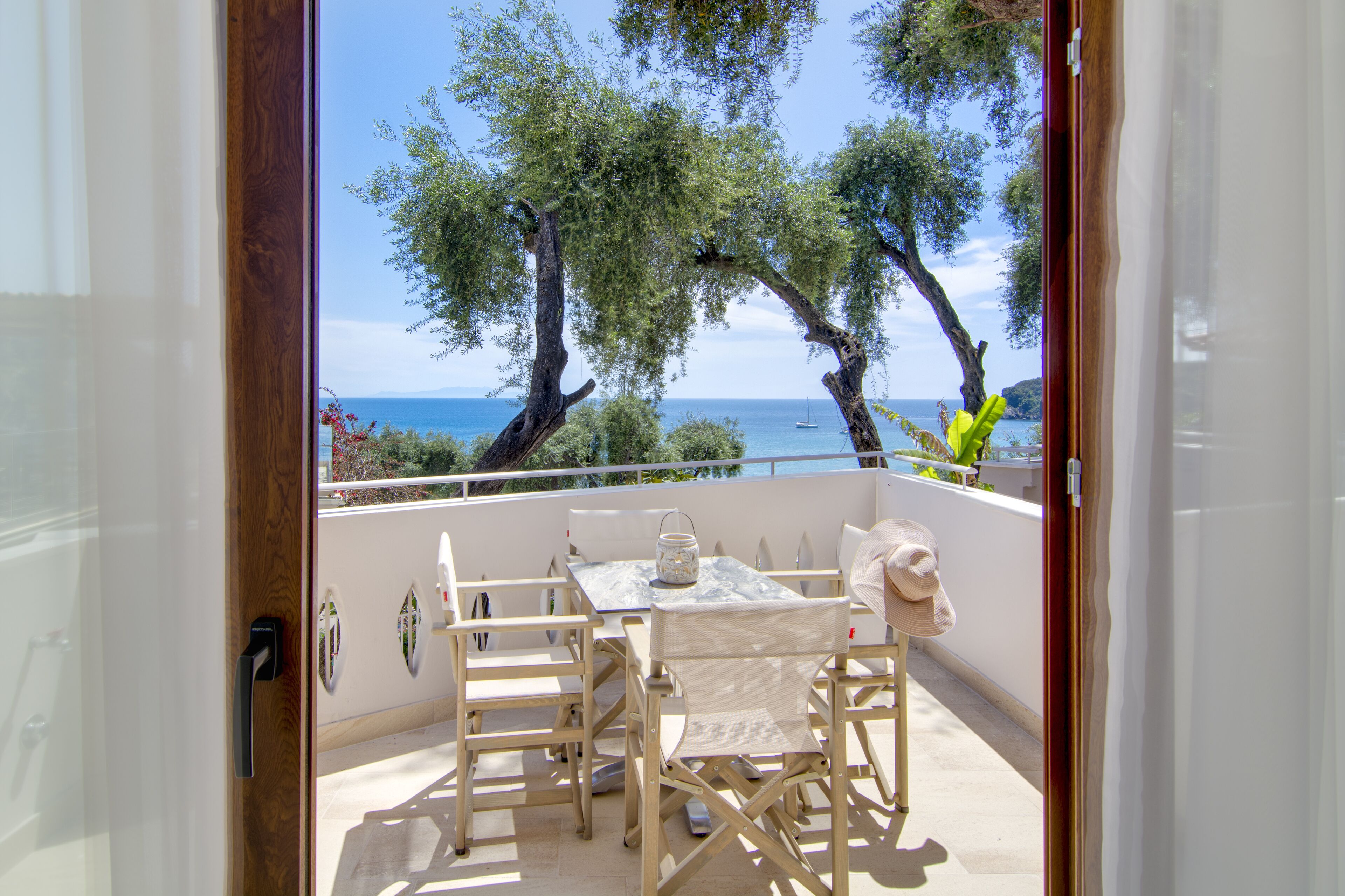 An open balcony with a table and chairs, overlooking a coastal area with trees and a boat in the distance.