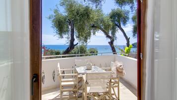 An open balcony with a table and chairs, overlooking a coastal area with trees and a boat in the distance.