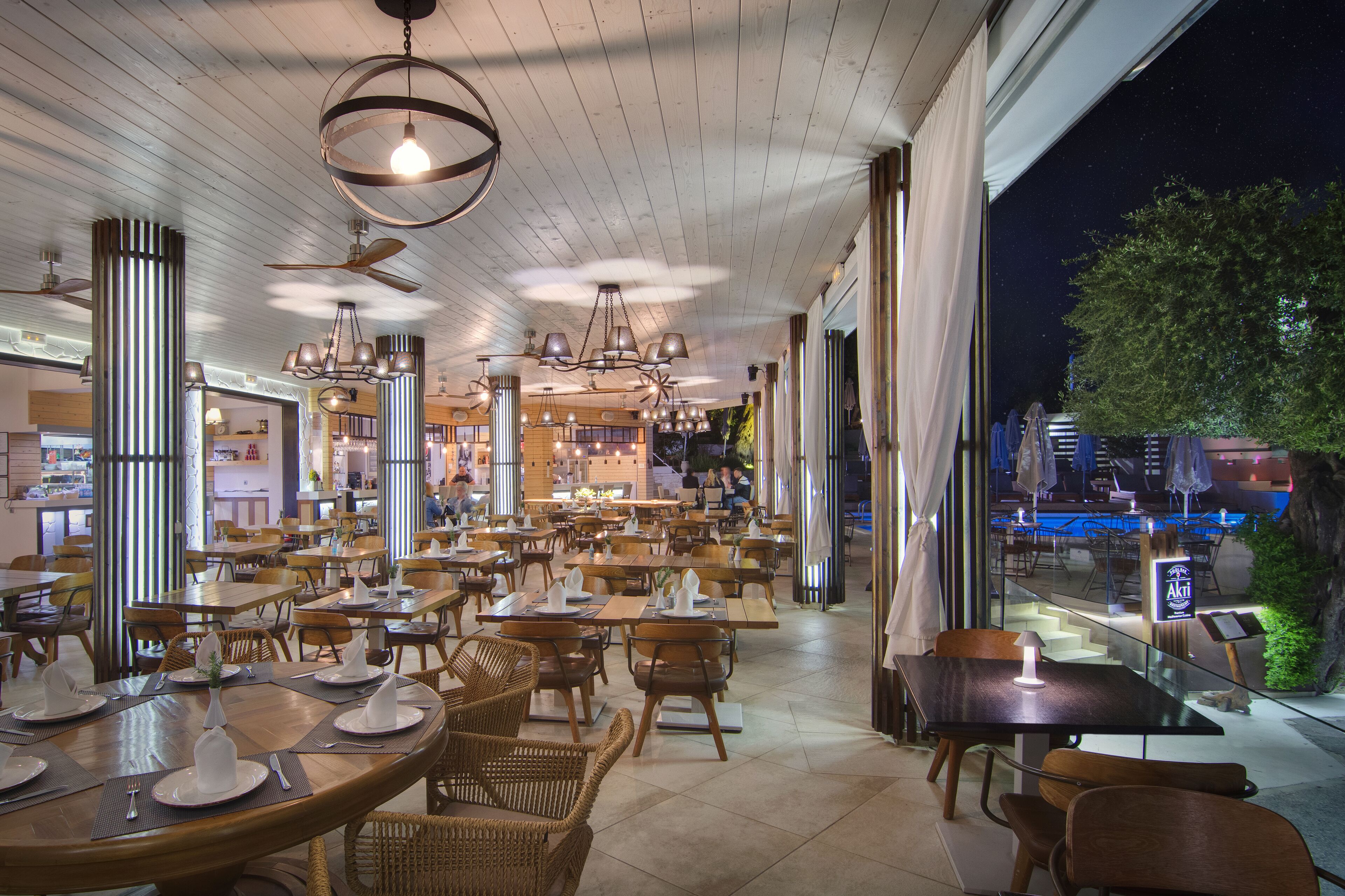 A spacious restaurant with multiple dining tables and chairs, large windows, and a view of the cityscape at night.