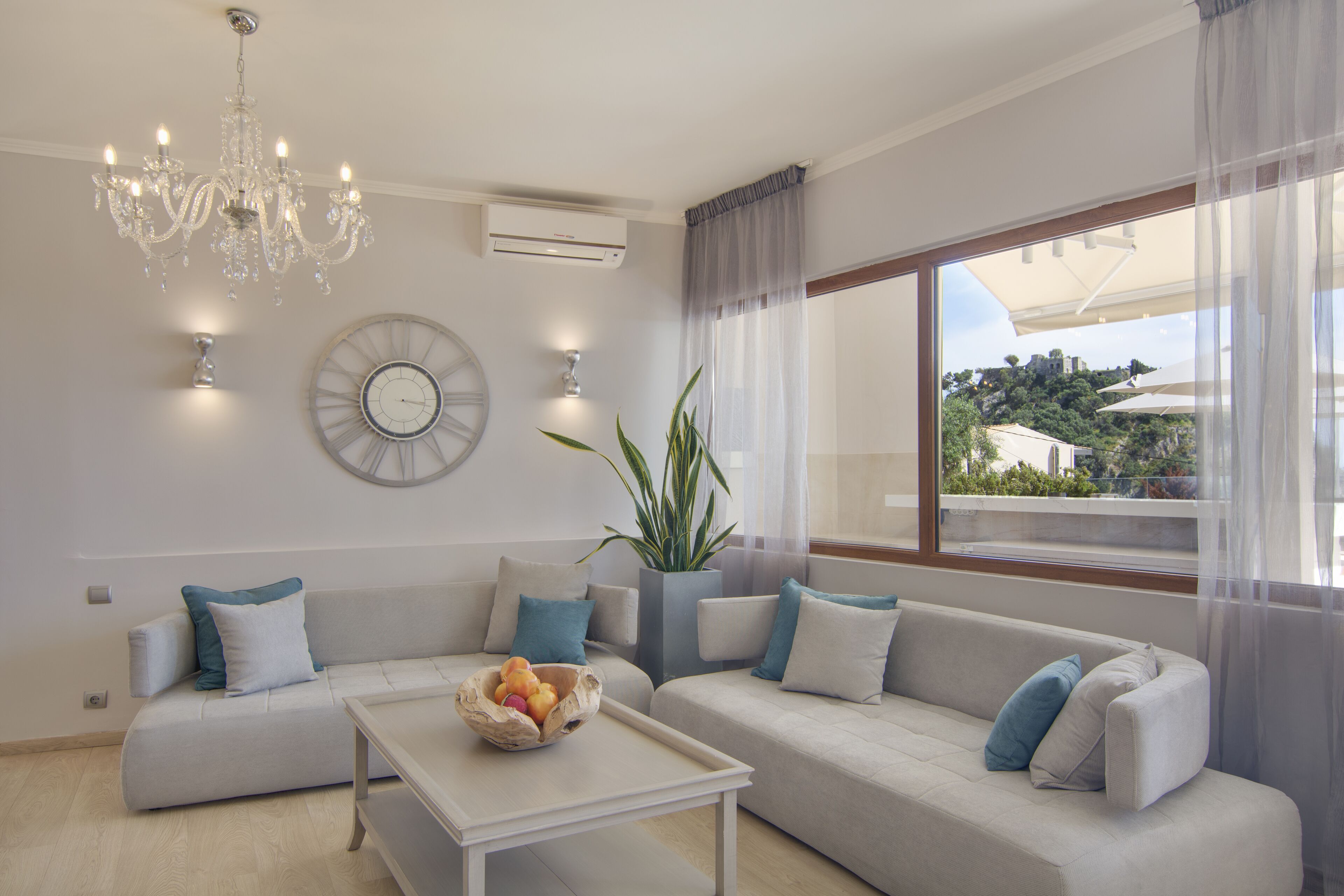 Una sala de estar moderna con una araña, un ventanal con vistas, un conjunto de sofás y una mesa de centro con un frutero.