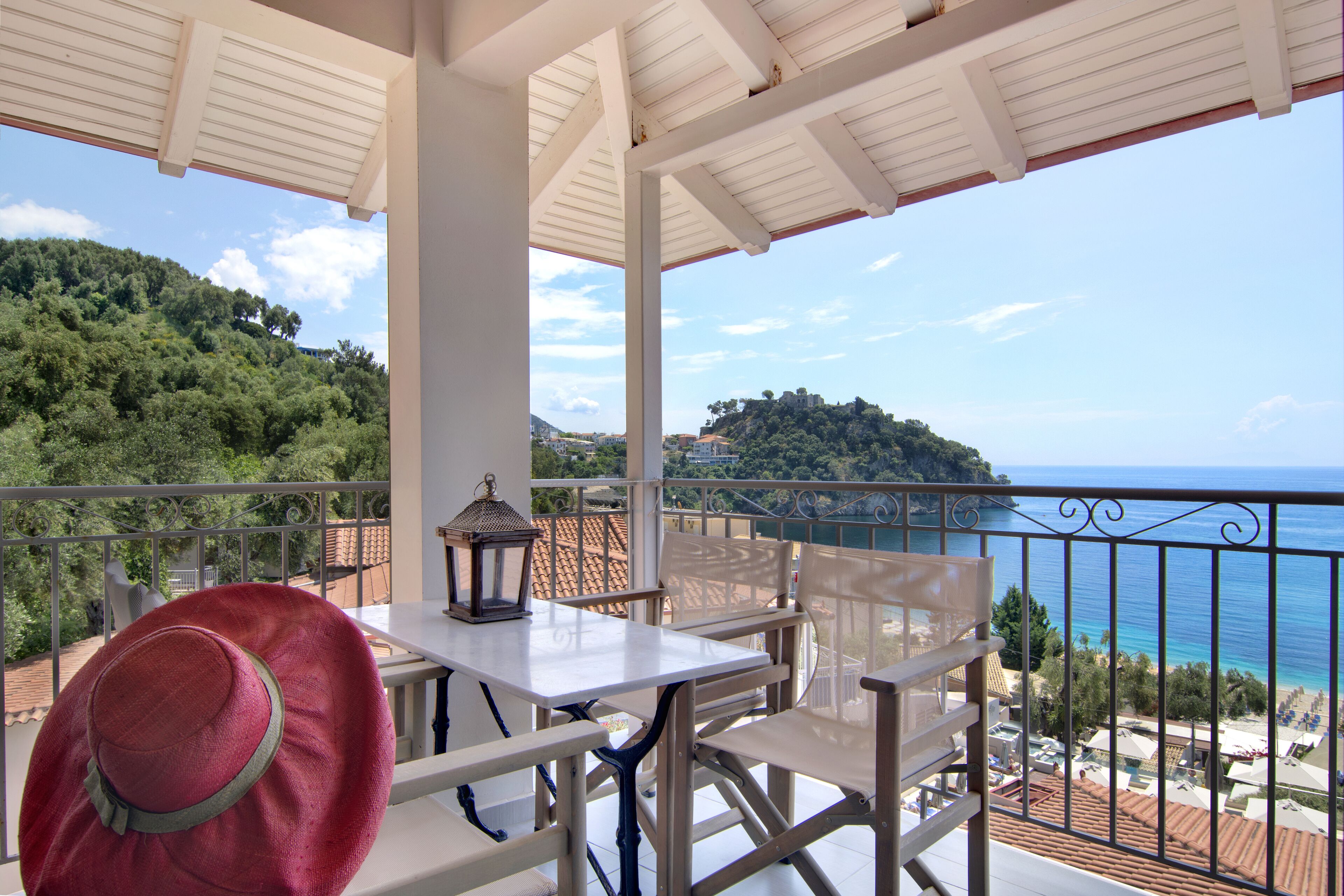 A balcony with a white table and chairs, overlooking a coastal view with trees and buildings.