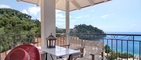 A balcony with a white table and chairs, overlooking a coastal view with trees and buildings.