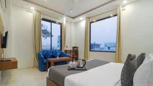 Habitación de hotel con cama, zona de estar con una silla y mesa, televisor montado en la pared y ventanas amplias con cortinas.