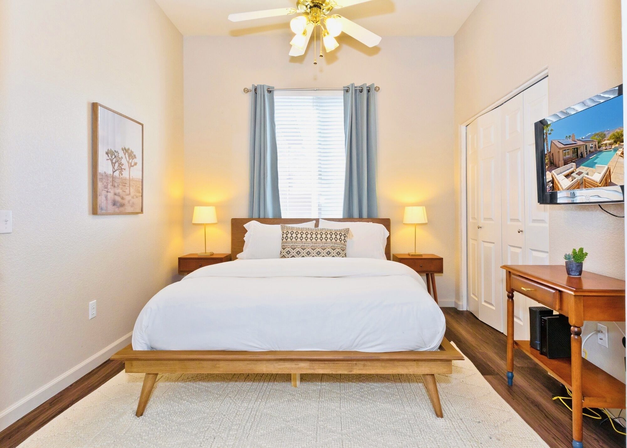 A bedroom with a bed, two bedside lamps, a ceiling fan, a TV on the wall, and a window with curtains.