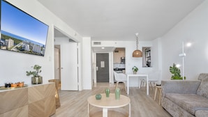 Ein modernes Wohnzimmer mit rundem Couchtisch, einer Sitzecke und einem an der Wand befestigten Fernseher. Im Hintergrund ist eine Küchenzeile erkennbar.
