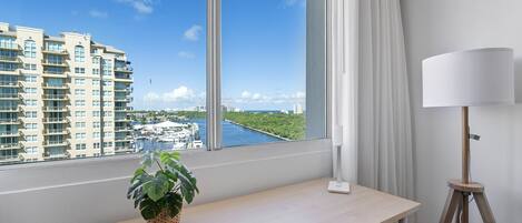 A hotel room with a large window offering a view of a marina and high-rise buildings.