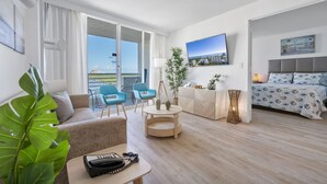 Ein modernes Hotelzimmer mit einer Couch, einem kleinen Tisch mit Telefon, einem Schreibtisch mit Lampe, einem Bett mit gemusterten Laken sowie einem Fernseher an der Wand und einem großen Fenster mit Blick auf die Stadt.