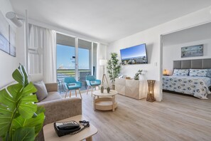 Et moderne hotelværelse med sofa, et lille bord med telefon, skrivebord med lampe, seng med mønstret sengetøj, fjernsyn på væggen og et stort vindue med udsigt over byen.