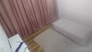 A small, sparsely furnished room with a white sofa, a wooden floor, and a pink curtain.