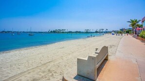 Ein Strand mit sandigem Ufer, einem klaren blauen Meer und einigen Booten. Eine Betonbank befindet sich auf der rechten Seite.