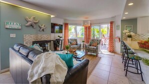 Ein Wohnzimmer mit einer Couch, Sesseln, einem Couchtisch und einem Fernseher. Vorhanden sind Vorhänge, ein Kamin und ein Essbereich mit Barhocker.