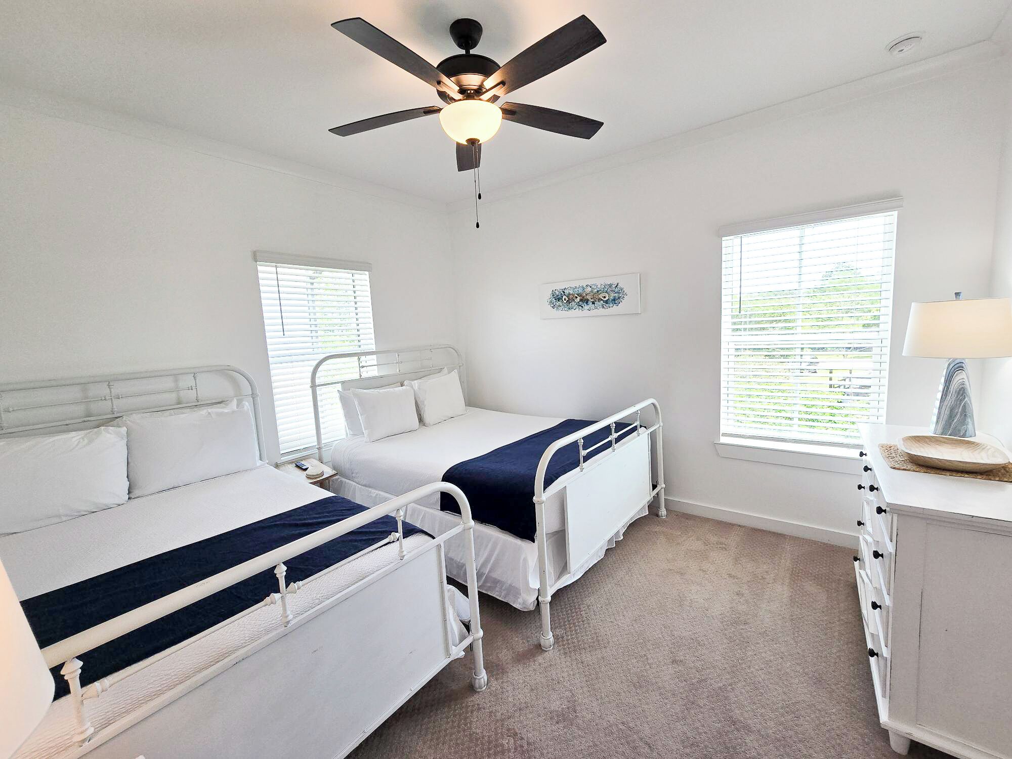 A bedroom with two beds, a ceiling fan, a bedside table with a lamp, and a window with blinds.