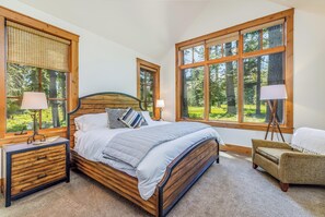 A bedroom with a wooden bed, two bedside tables, a chair, and large windows with a view of trees.