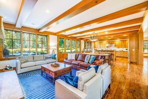 A spacious living area with a large sofa, a coffee table, and a fireplace. There are large windows providing natural light and a view of the outdoors.