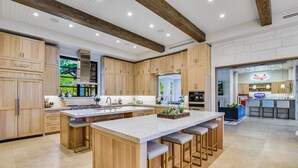 A spacious kitchen with wooden cabinets, a large island with bar stools, and a view of the outdoor area. - Above Lakeside I Lakefront | Pickleball I Pool/Spa l Firepit I Luxe Services (Austin)